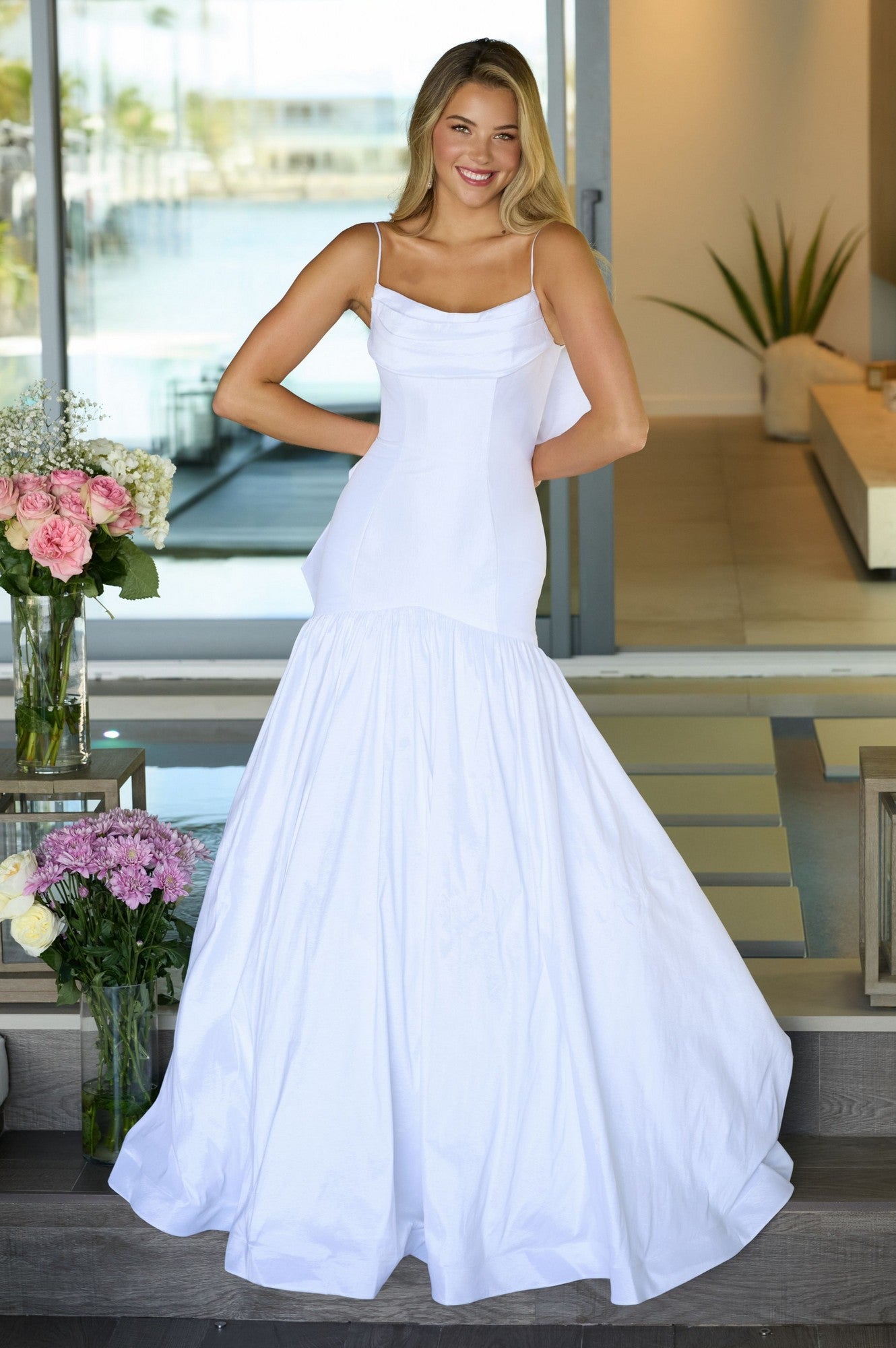 A woman stands indoors smiling with hands on hips, wearing the Ava Presley 26422 Drop-Waist Mermaid Prom Gown. Flowers and modern decor accent the background.